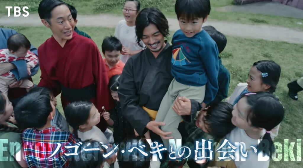 TBSの放送画面。画面下部に「Encounter with Nogoon Beki」という英語の透かし文字の上に、日本語で「ノゴーン・ベキとの出会い」というテロップが表示されている。中央には黒い衣装を着た若き日のノゴーン・ベキがおり、青い服の男の子を笑顔で抱きかかえている。左側には赤い衣装を着た若きバトラカが、子どもたちを見つめて穏やかに微笑んで立っている。彼らの周りには大勢の子どもたちが集まり、皆で和やかに笑い合っている屋外の草地の風景。