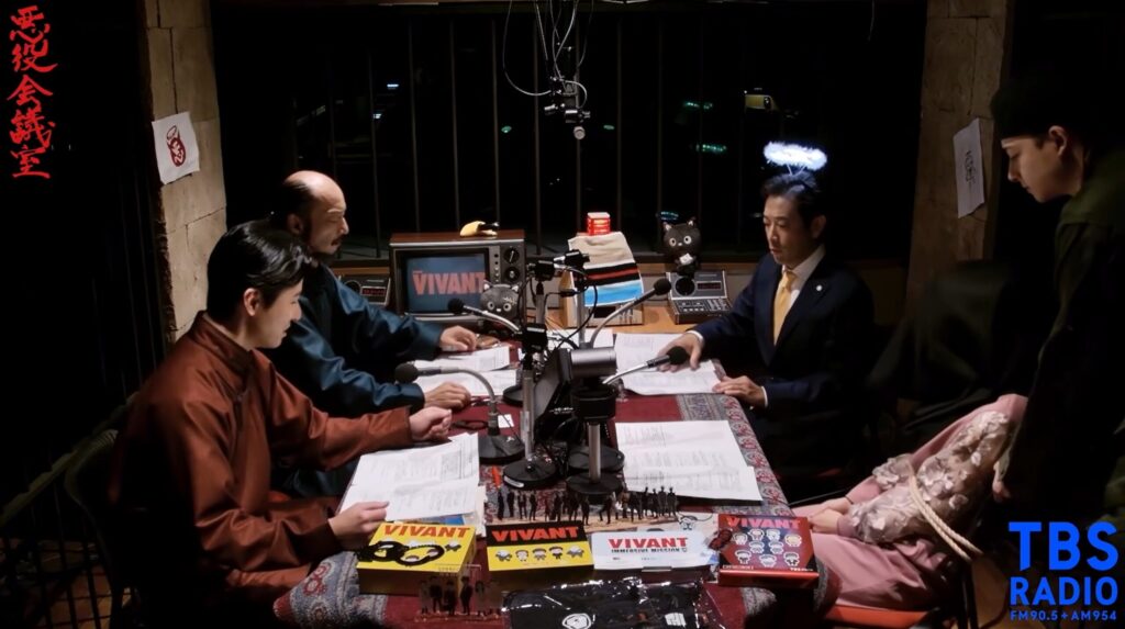 TBSラジオ『VIVANT 悪役会議室』の収録風景です。薄暗い部屋で、テーブルを囲みマイクに向かう迫田孝也さんら出演者の姿が映っています。机にはVIVANTの台本やグッズが並んでいます。