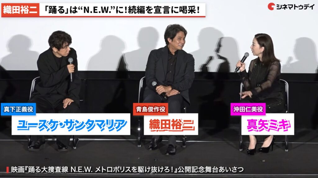 映画『踊る大捜査線』の舞台あいさつで、ユースケ・サンタマリアさん、織田裕二さん、真矢ミキさんが椅子に座って和やかに語り合う様子です。画面上部には織田さんの続編宣言を伝えるテロップが表示されています。