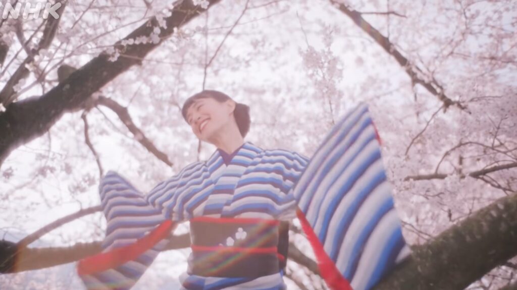 満開の桜の木の下で、青と白の鮮やかなストライプ柄の着物を着た若い女性が、両手を広げて晴れやかな笑顔を見せている場面です。女性の黒い帯には可愛らしい白い花の模様があしらわれています。画面いっぱいに淡いピンク色の桜の花と枝が広がり、ふんわりとした温かく希望に満ちた春の空気が感じられます。左上には薄くNHKのロゴが配置されており、連続テレビ小説のオープニングらしい明るい雰囲気が伝わる一枚です。
