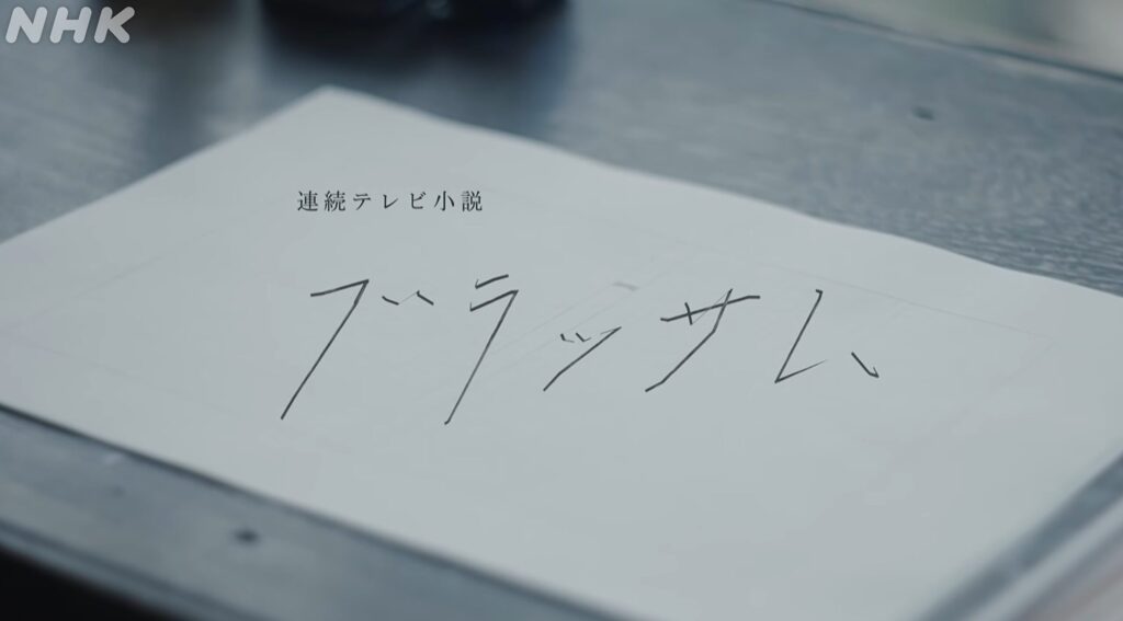 薄暗い机の上に置かれた一枚の白い紙がクローズアップされた画像です。紙の上部には小さく「連続テレビ小説」と印字されており、その下には伸びやかな手書き風の文字で大きく「ブラッサム」とタイトルが書かれています。画面の左上には白い文字でNHKのロゴが配置されています。シンプルでありながら、これから始まる物語への期待を感じさせるような、静かで落ち着いた雰囲気のタイトルバックのワンシーンです。