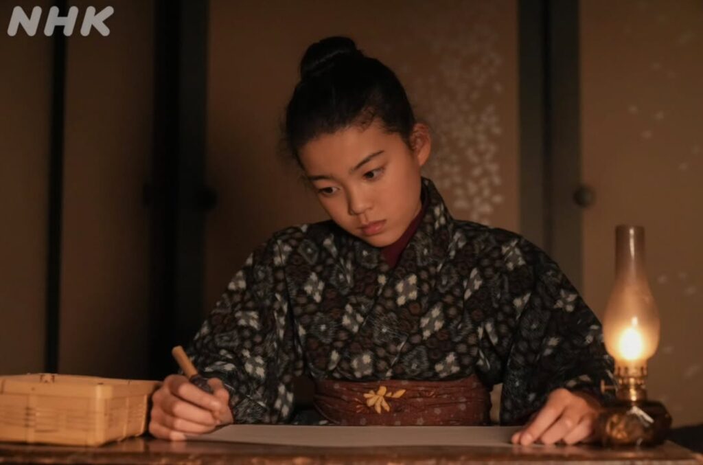 薄暗い部屋の中、ランプの温かい明かりを頼りに、村上蘭さんが木の机に向かっている場面です。髪を結い上げ、模様のある着物を着た彼女は、右手に筆記用具を持ち、真剣な眼差しで紙を見つめながら書き物を行っています。画面左上にはNHKのロゴがあります。