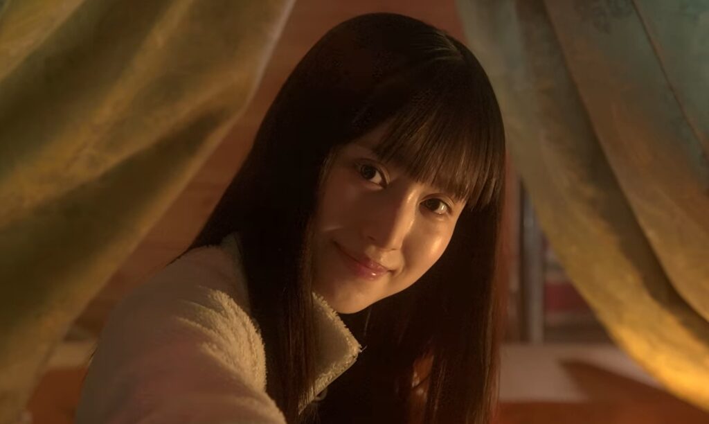 映画『室井慎次 敗れざる者』に登場する日向杏。テントの幕間から顔を出し、黒髪ストレートで微笑みを浮かべながらカメラを見つめるシーン。あたたかい照明のもと、どこか謎めいた表情を見せるワンシーン。