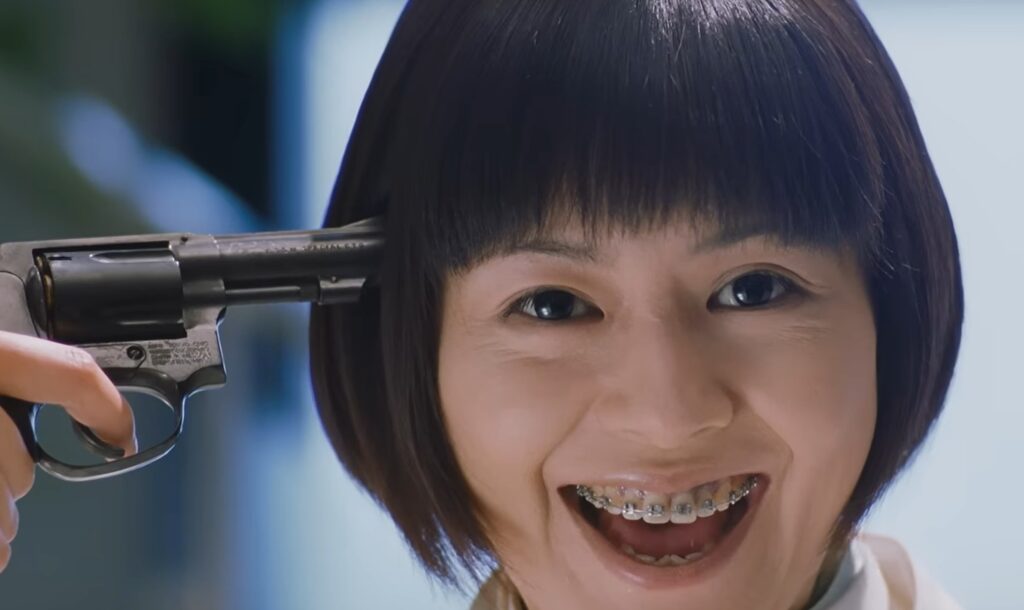 映画『室井慎次 敗れざる者』の回想シーン。小泉今日子演じる日向真奈美が、ボブヘアに歯科矯正器具をつけた姿で狂気的な笑顔を浮かべる。右手に持った黒いリボルバー拳銃を自身の右こめかみに突きつけている、緊迫したワンシーン。