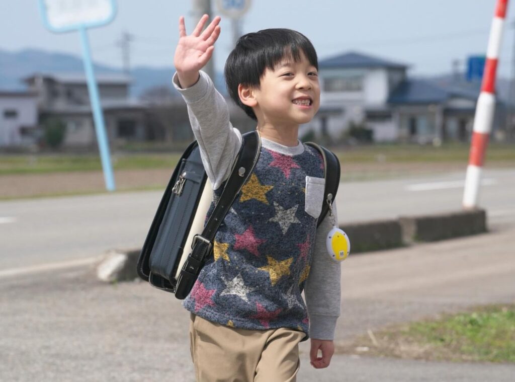 映画『室井慎次 敗れざる者』のワンシーンです。黒いランドセルを背負い、星柄の服を着た小学生の男の子（リクさん）が、明るい笑顔で右手を高く挙げて手を振っています。背景には見通しの良い道路と住宅街が広がり、元気いっぱいに歩いている様子です。