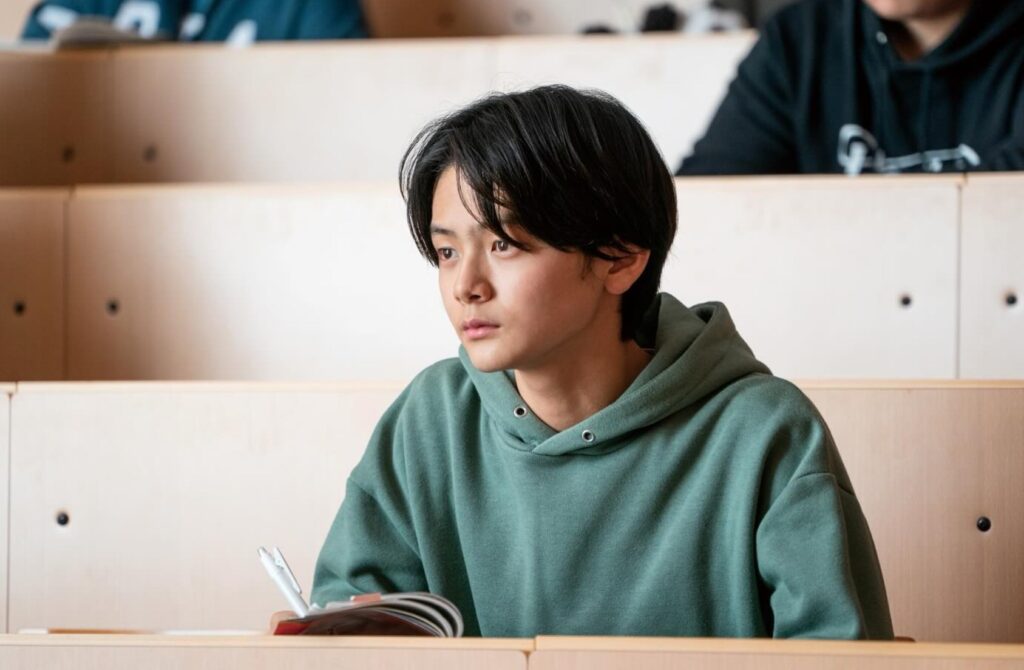 映画『室井慎次 敗れざる者』のワンシーン。タクを演じる若い男性が、緑色のパーカーを着用し、講堂の木製デスクに座っている。ペンとノートを手に、真剣な眼差しで左を見ている。他の学生の姿も背景にあり、集中した講義風景。