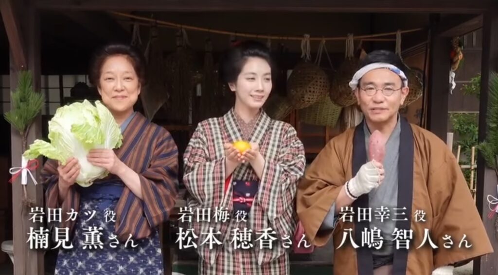 朝ドラ『ブラッサム』のワンシーンです。左から、白菜を手にした岩田カツ役の楠見薫さん、みかんを持つ岩田梅役の松本穂香さん、さつまいもを持つ岩田幸三役の八嶋智人さんが並んで微笑んでいます。古い日本家屋を背景に、3人の和やかな雰囲気が伝わる一枚です。