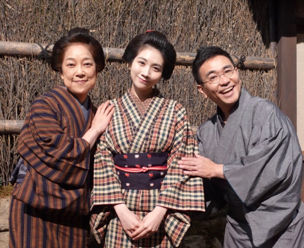 朝ドラ『ブラッサム』のオフショット。和服姿の楠見薫さん、松本穂香さん、八嶋智人さんの3人が並んで微笑んでいます。楠見さんと八嶋さんが松本さんの肩に手を添え、親密な家族の雰囲気が伝わる一枚。背景は茅葺き壁です。