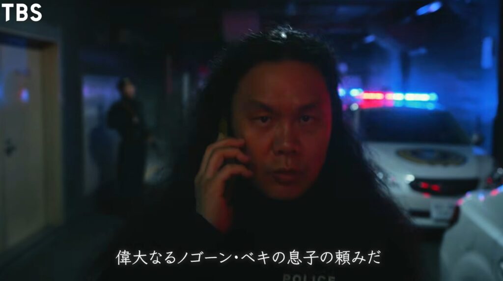 TBSドラマ『チンギス』の場面画像です。夜の屋外でパトランプが光る警察車両を背に、長髪の男性が真剣な眼差しでスマートフォンを耳に当て通話しています。画面下には「偉大なるノゴーン・ベキの息子の頼みだ」という字幕が表示されています。