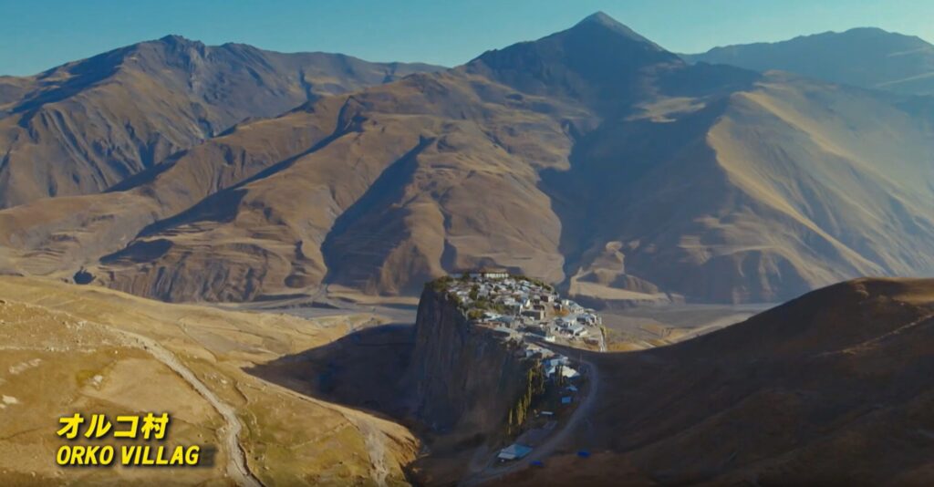 ドラマ『VIVANT』に登場するバルカ共和国のオルコ村の全景です。荒涼とした壮大な山々に囲まれ、切り立った断崖絶壁の上に小さな集落が広がっています。画面左下には「オルコ村 ORKO VILLAG」という黄色いテロップが表示されています。