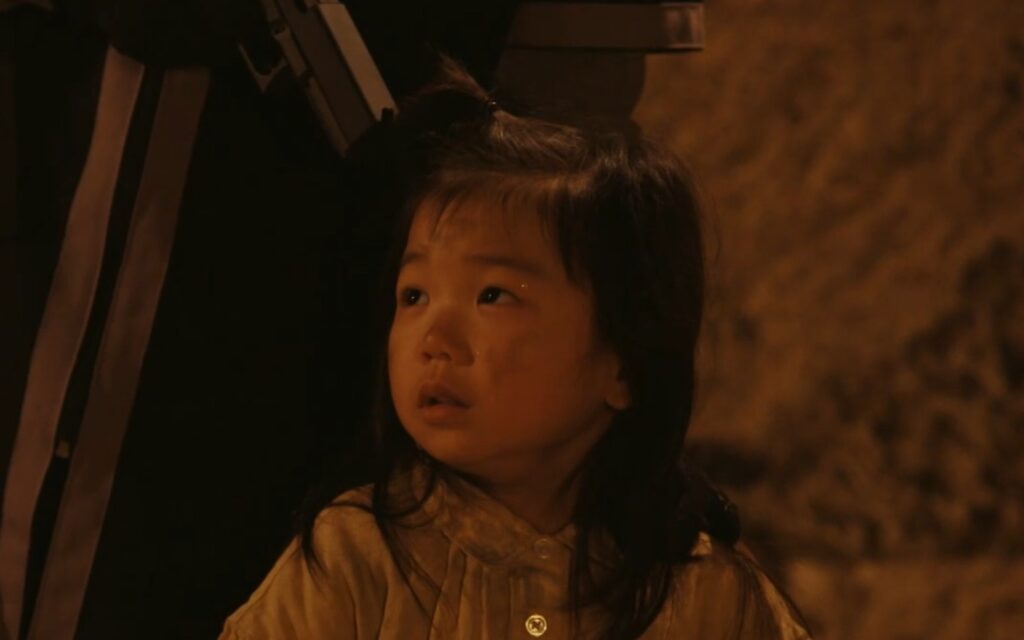 ドラマ『VIVANT』第19話より、核融合技術者エリシャの幼い娘・プリシャのアップ写真。薄暗い中で顔に薄汚れを浮かべ、不安と戸惑いが入り混じった瞳でじっと上を見つめており、監獄内の緊迫した状況と幼い彼女の情景が浮かぶ印象的な一枚です。
