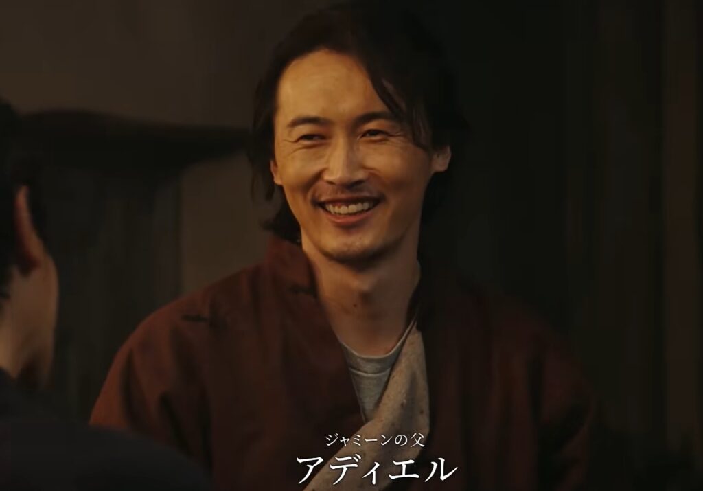 ドラマ『VIVANT』より、ジャミーンの父アディエルの登場シーン。茶色の衣装を纏い、相手に向かって優しく温かい笑顔を浮かべています。画面下部には「ジャミーンの父 アディエル」とテロップが表示されており、彼の誠実な人柄がにじみ出る印象的な一枚です。