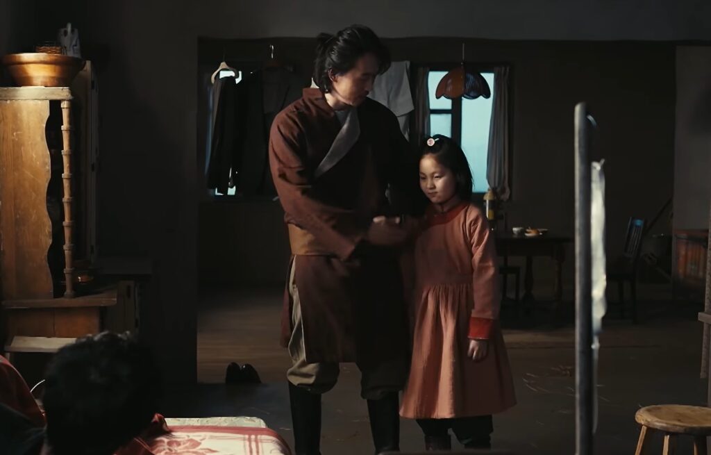 ドラマ『VIVANT』より、父アディエルと娘ジャミーンのワンシーン。落ち着いた雰囲気の室内で、茶色の民族衣装を着たアディエルが愛娘ジャミーンの身なりを整えています。ピンク色の衣装を纏うジャミーンとの、温かい親子の絆が伝わる一枚です。