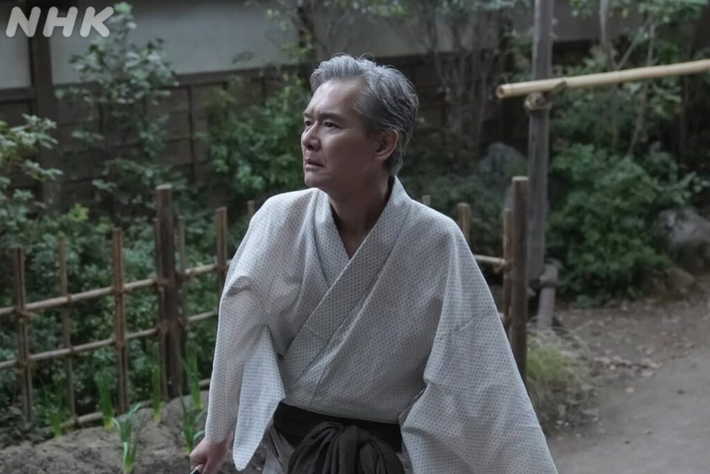 NHKのブラッサム　清治（きよじ）の前に現れた「もう一人の清治」のワンシーン。白髪で伝統的な和装（白い着物）に身を包んだ年配の男性が、厳しい表情で庭園の左側を睨んでいる。背景には竹垣や植物があり、緊張感が漂う。