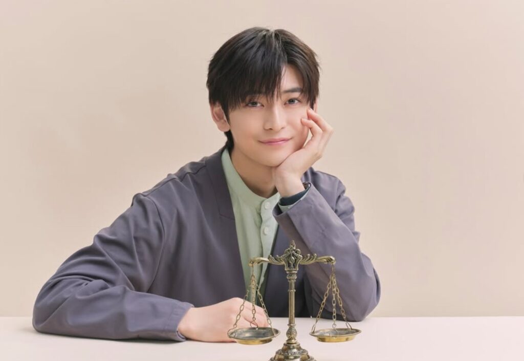 ドラマ『南田南弁護士は天才肌』で船井湊人を演じる高橋文哉さんが、机の上の天秤を前にして、左手でほおを突いて優しく微笑んでいる。グレーのジャケットを着用。シンプルな背景。
