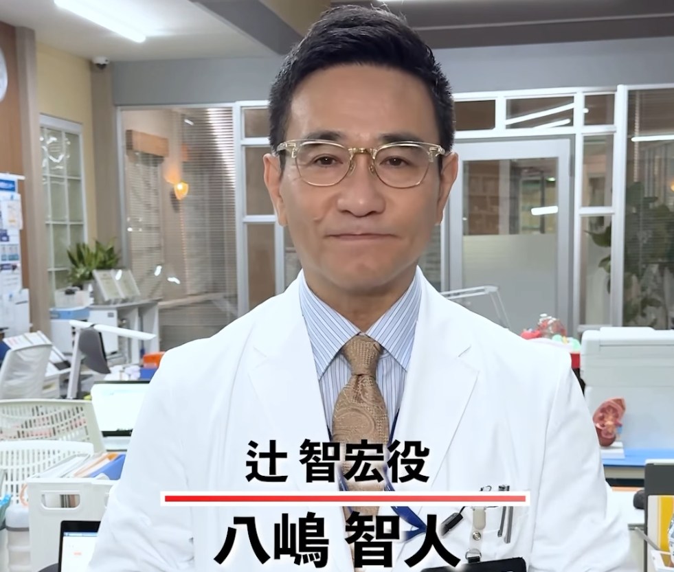 病院の診察室を背景に、ドラマ『救命病棟24時』で辻智宏役を演じる俳優の八嶋智人さんが白衣とメガネを着用し、正面を見据えて立っているシーン。画面下部には「辻智宏役 八嶋智人さん」のテロップが表示されている。