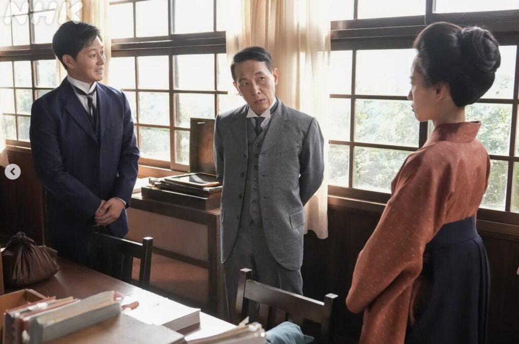 NHKドラマ『ブラッサム』の一場面。レトロな職員室で、グレーの3ピーススーツを着た厳しい表情の上野教頭が、化粧のことで珠(和服姿)を指摘している。珠は教頭と向き合っており、左にはネイビーのスーツの男性が立っている。緊張した対話の様子。