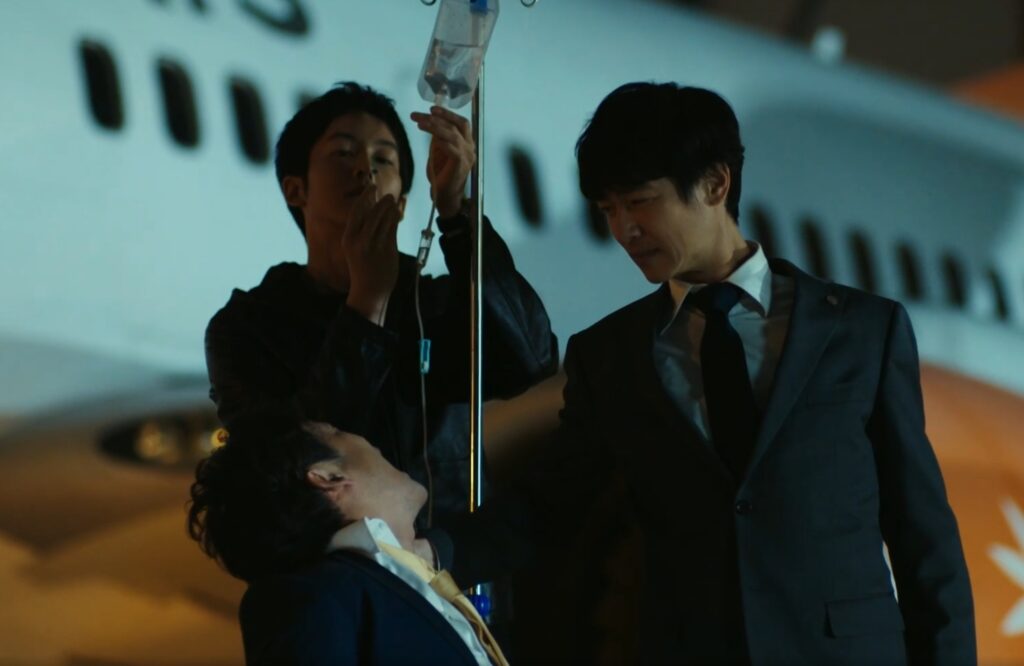 『VIVANT』第4話、夜の滑走路。黒須(松坂桃李)が点滴を準備する傍らで、乃木(F、堺雅人)が拘束された山本(迫田孝也)を冷徹な表情で見下ろし尋問している。背景には飛行機の機体が写っている。