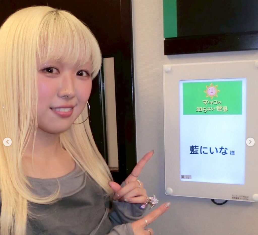 金髪の女性が笑顔で、番組名「マツコの知らない世界」と自身の名前「藍にいな 様」が書かれたパネルを指さしている。YOASOBIのMVなどを手がける藍にいなさんが、番組出演の際に楽屋前で撮影したオフショット。