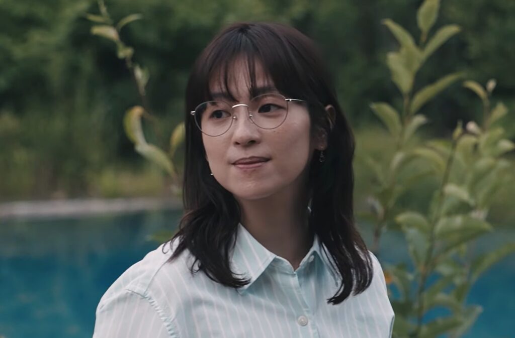 ドラマ「LOST10(ロストテン)」で亜矢を演じる中村アンさんが、自然の中で眼鏡をかけ、水色のストライプシャツ姿で佇むシーン。穏やかな表情で、左方向を見つめている様子。