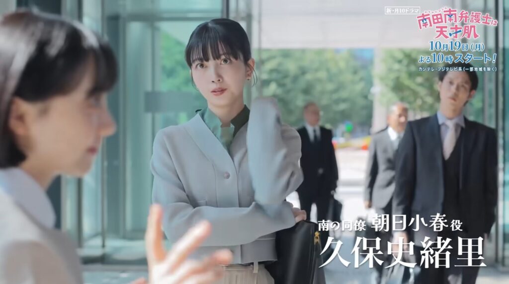 ドラマ「南田南弁護士は天才肌」の朝日小春役、久保史緒里さんが、グレーのジャケット姿でビルのエントランスに立つ。髪に手をやり、まっすぐな視線で手前の人物と向き合う。背景にはスーツ姿の男性たちが歩く。画面にはドラマのロゴと彼女の名前。