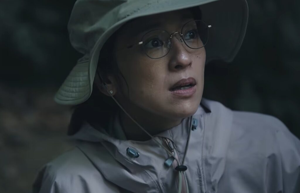 ドラマ『LOST10』で中村アンさん演じる、登山ツアーで遭難し保険金をめぐる疑惑が持たれる亜矢のシーン。メガネをかけアウトドア用のハットとウェアを着用し、薄暗い森の中で切迫した表情で何かを見上げる様子。