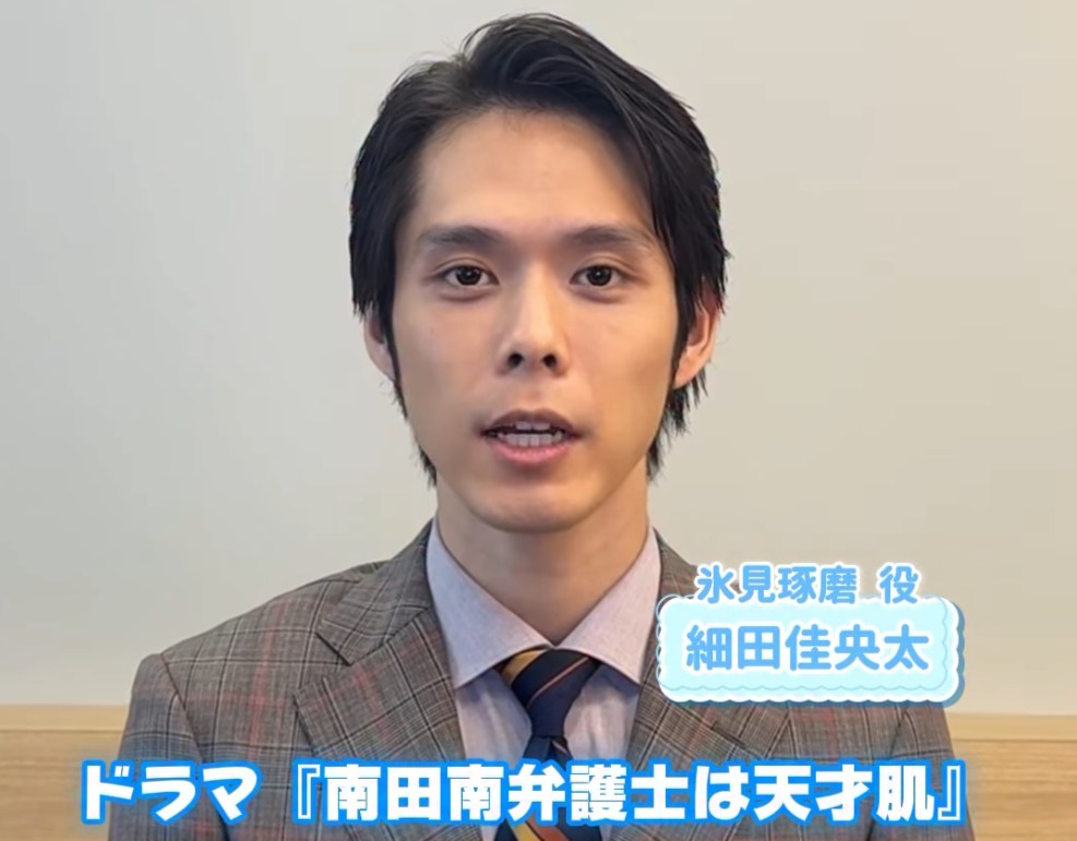ドラマ『南田南弁護士は天才肌』の撮影現場。氷見琢磨役を演じる俳優・細田佳央太が、チェック柄のスーツにネクタイ姿で、カメラに向かって真剣な表情で話している様子を捉えたポートレート。