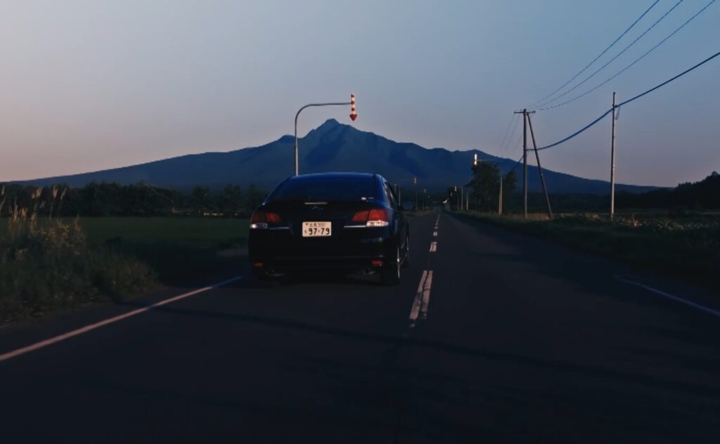 夕暮れのまっすぐな道路を手前の車が走行している様子。左手前には赤と白の縞模様の下向き矢印の形をした矢羽根（固定式視線誘導柱）が設置されており、遠くには大きな山がそびえています。