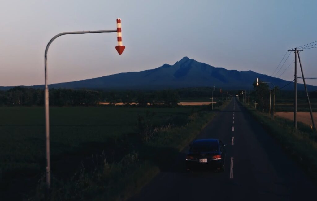 夕暮れのまっすぐな道路を手前の車が走行している様子。左手前には赤と白の縞模様の下向き矢印の形をした矢羽根（固定式視線誘導柱）が設置されており、遠くには大きな山がそびえています。