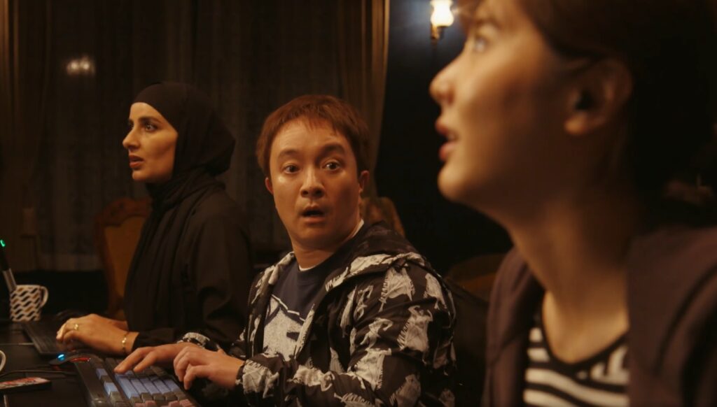 暗い室内で、3人の人物が驚いた表情で画面右を注視している。中央のプリントジャケットの男性と黒いヒジャブを着用した女性はキーボードの前に座り、全員が何かに凝視している劇的な瞬間。