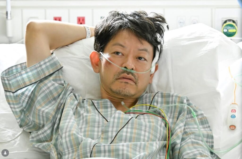 『救命病棟24時』より、山中崇さんが演じる小野川卓が、チェック柄の患者衣姿で病院のベッドに横たわり、鼻カニューレとモニターコードをつけて、虚ろな表情で横たわっているシーン。