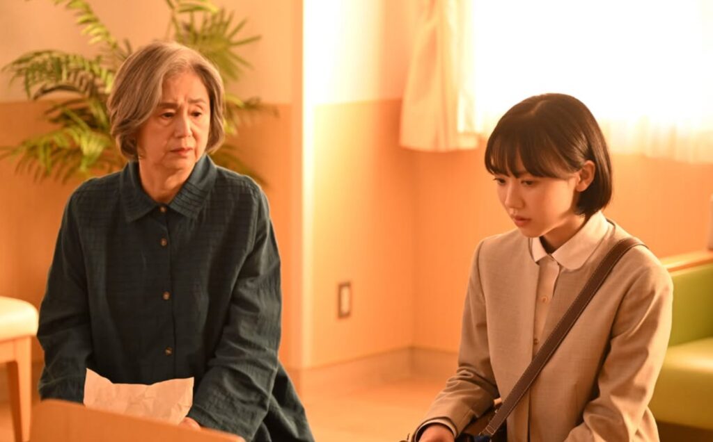ドラマ『南田南弁護士は天才肌』第1話、一ノ瀬圭子役の朝加真由美。南田南弁護士のとなりに座り、心配そうな、悲しげな表情で前を見つめている。
