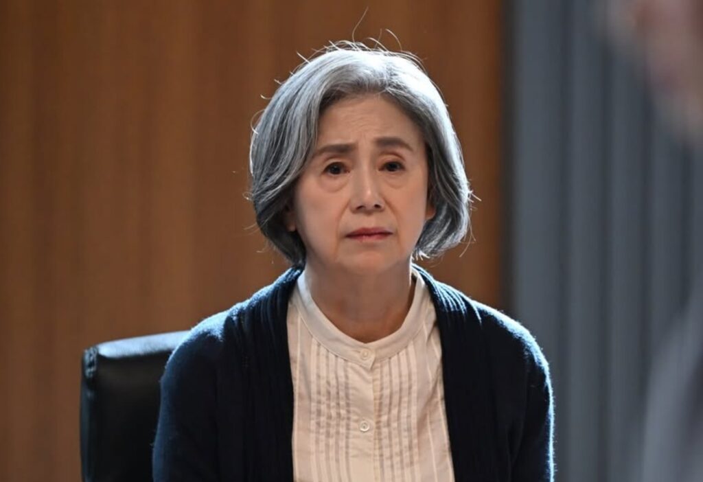 ドラマ『南田南弁護士は天才肌』第1話、一ノ瀬圭子役の朝加真由美。黒いオフィスチェアに座り、心配そうな、悲しげな表情で前を見つめている。