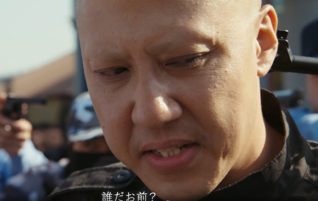 ドラマ『VIVANT』でスキンヘッド姿の周浩然を演じるリンリンさんが、周囲に銃口が向けられる中、険しい表情を浮かべて「誰だお前？」と語りかける緊迫したワンシーン。
