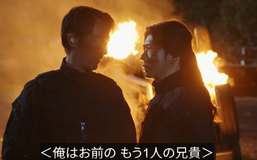 ドラマ『VIVANT』のワンシーン。激しい炎が燃え上がる夜の野外で、黒い服を着たノコルと乃木が至近距離で向き合い、緊張感のある眼差しを交わしている様子。画面下部には「＜俺はお前の もう1人の兄貴＞」と字幕が表示されている。