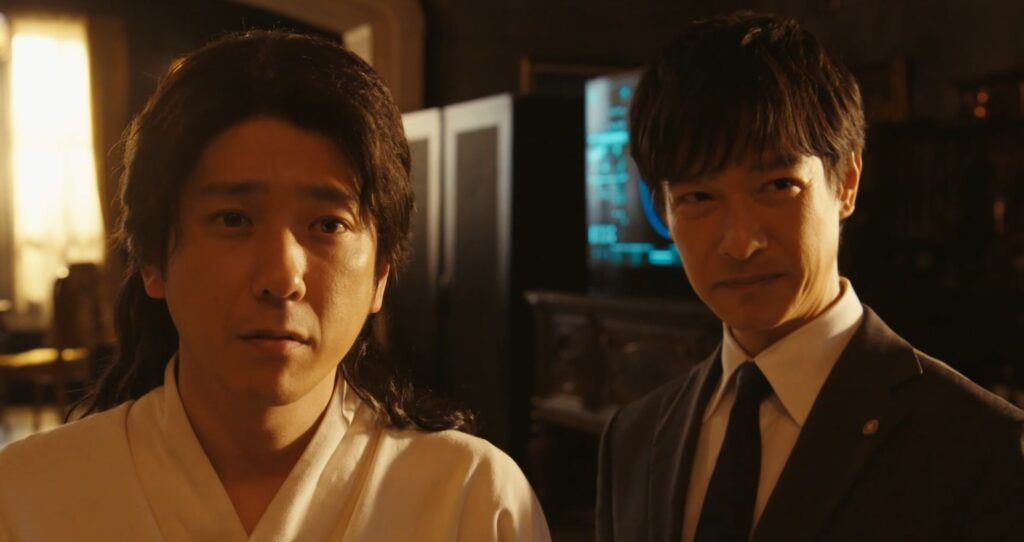 ドラマ『VIVANT』のワンシーン。薄暗い室内で並んで立つ乃木憂助とノコル。スーツ姿の乃木が微かに笑みを浮かべ、白い民族衣装を着たノコルが真剣な表情で正面を見つめている姿。