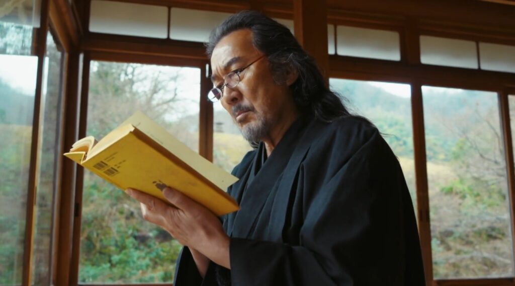 ドラマ『VIVANT』のワンシーン。和服姿で眼鏡をかけたノゴーン・ベキが、日本家屋の窓辺で黄色い表紙の本を手に持ち、真剣な表情でページを見つめているアップのカット。