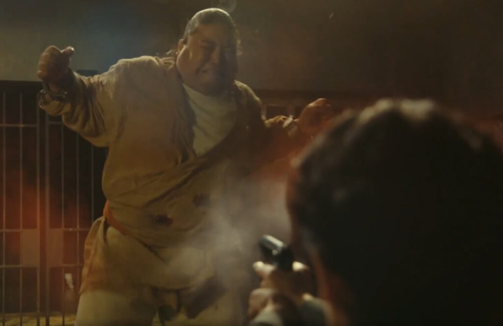 ドラマ『VIVANT』のワンシーン。手前で銃を構えるリュウ・ミンシュエンと、発砲時の煙が漂う中で痛みに耐えるように体を強張らせるドラムの緊迫した場面。