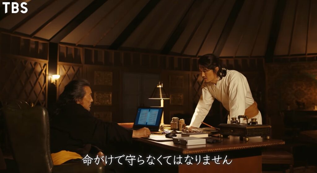 TBSのドラマ『VIVANT』のワンシーン。ゲルの中の重厚なデスクに向かい合って座るベキと、寄り添うように立つアディエルが真剣な表情で話し合っており、「命がけて守らなくてはなりません」という字幕が表示されている。
