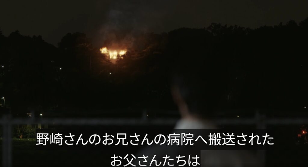 ドラマ『VIVANT』より、暗闇のなか激しく炎を上げて燃え上がる山中の建物を手前から人物が見つめている回想シーン。「野崎さんのお兄さんの病院へ搬送されたお父さんたちは」というテロップが表示されている。