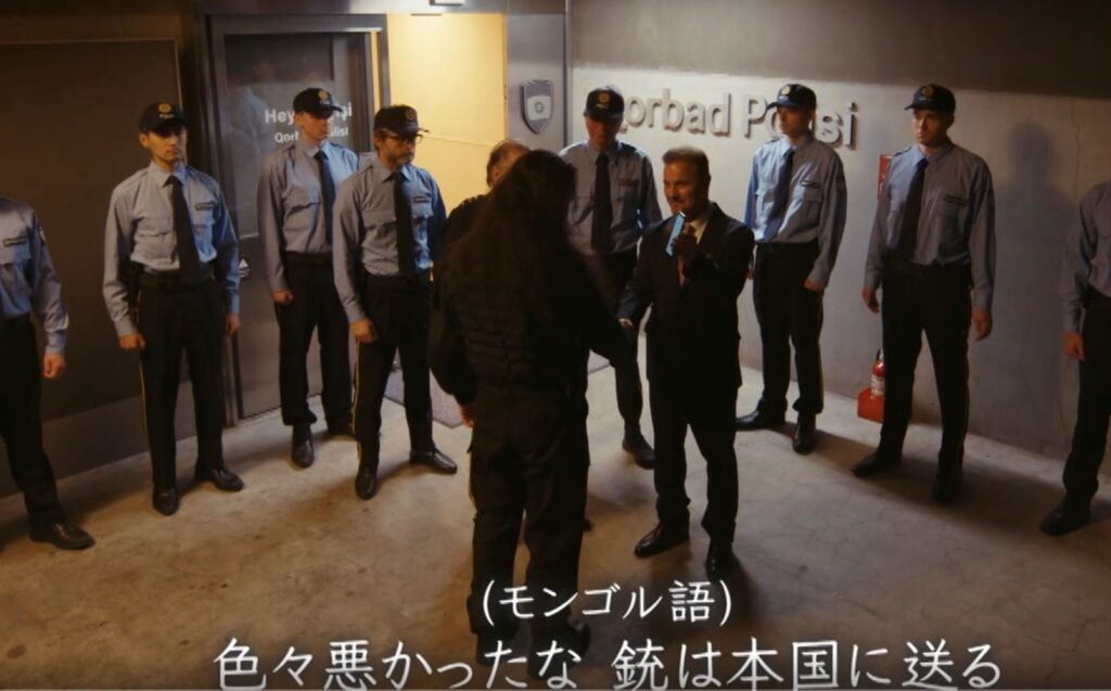 『VIVANT』第22話のワンシーン。ゴルバド警察に一時捕まったチンギスが、警察官たちに取り囲まれる中、スーツ姿の男性と握手を交わして釈放され、自由の身となる場面。