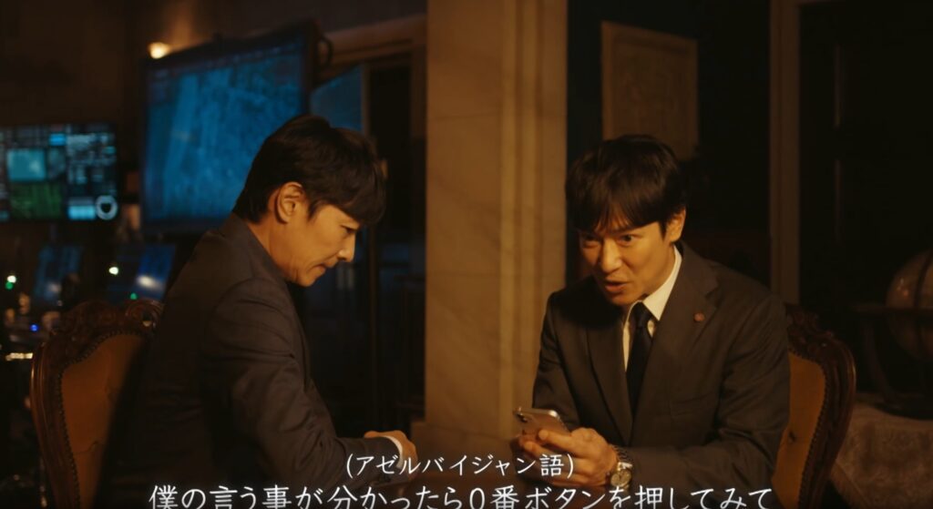 『VIVANT』のワンシーン。スーツ姿の乃木憂助がスマートフォンを手に持ち、アゼルバイジャン語で「僕の言う事が分かったら0番ボタンを押してみて」と真剣な表情で話しかけている様子。