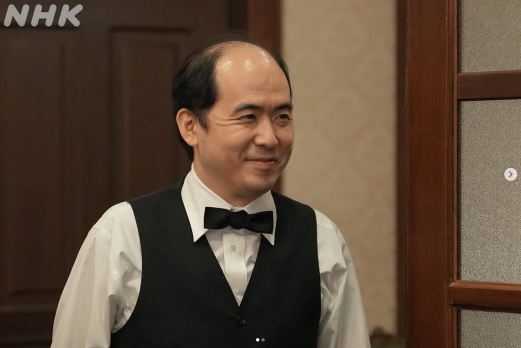 NHKの連続テレビ小説『ブラッサム』で西洋レストラン「悠樂軒」の店長・熊田を演じる斎藤司さん。白いシャツに黒いベスト、蝶ネクタイを着用し、穏やかな笑みを浮かべているワンシーン。