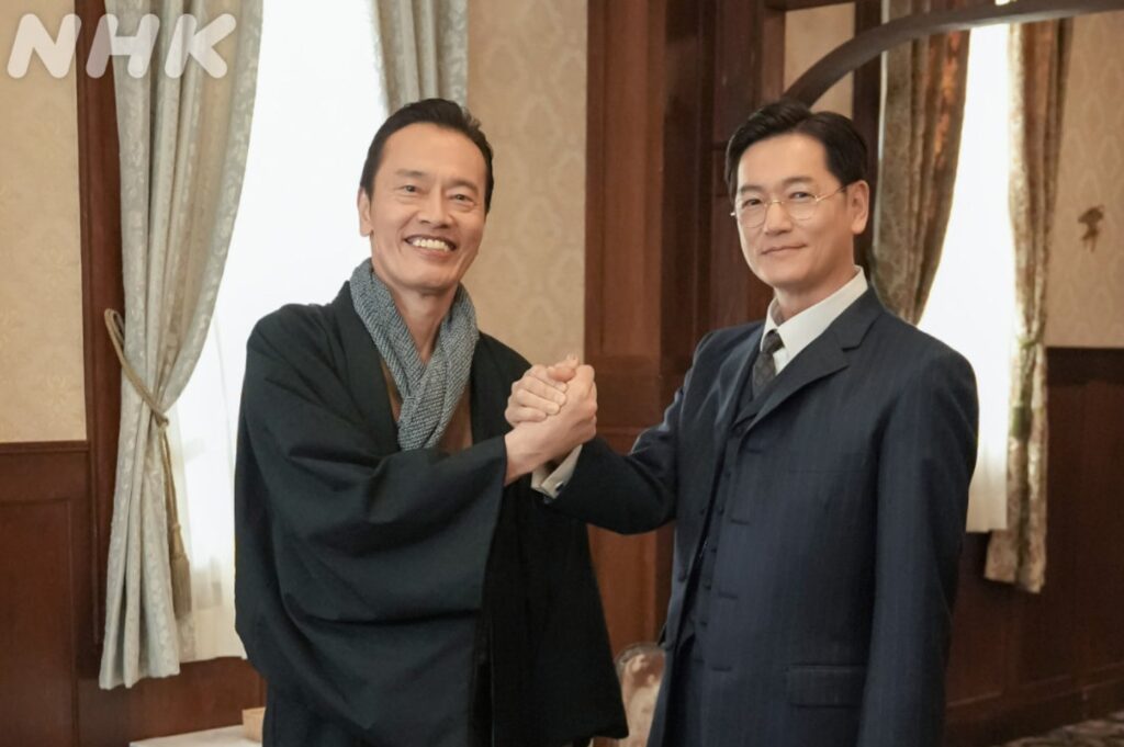 ドラマ『ブラッサム』のワンシーン。着物姿の黒崎大三郎を演じる遠藤憲一と、スーツ姿で眼鏡をかけた山中勇三を演じる井浦新が、笑顔で固く握手を交わしている場面。