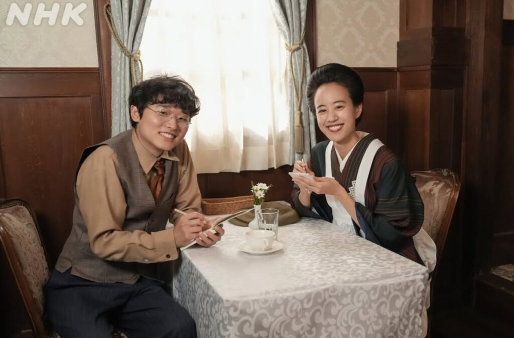 『ブラッサム』のワンシーン。レトロな喫茶店のテーブルに並んで座り、ノートとペンを手にカメラへ笑顔を向ける、左良太役の北野秀気さんと和服姿の女性。