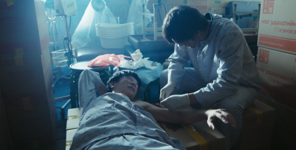 ドラマ『VIVANT』第1シーズン第1話で、病院の物置のような場所で痛みに耐えながら横たわる乃木と、その横にかがみ込み、手袋をはめて乃木の腕の傷口を真剣な表情で縫合処置する野崎の姿をとらえたワンシーン。